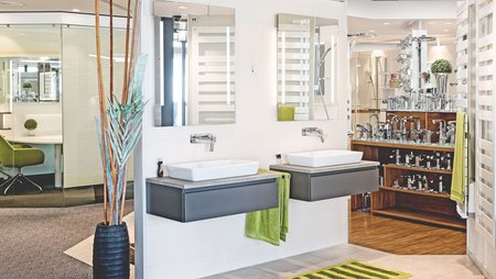 Helles Badstudio mit zwei wandhängenden Waschtischen, großen Spiegeln, grünen Handtüchern und umfangreicher Armaturenausstellung im Hintergrund.